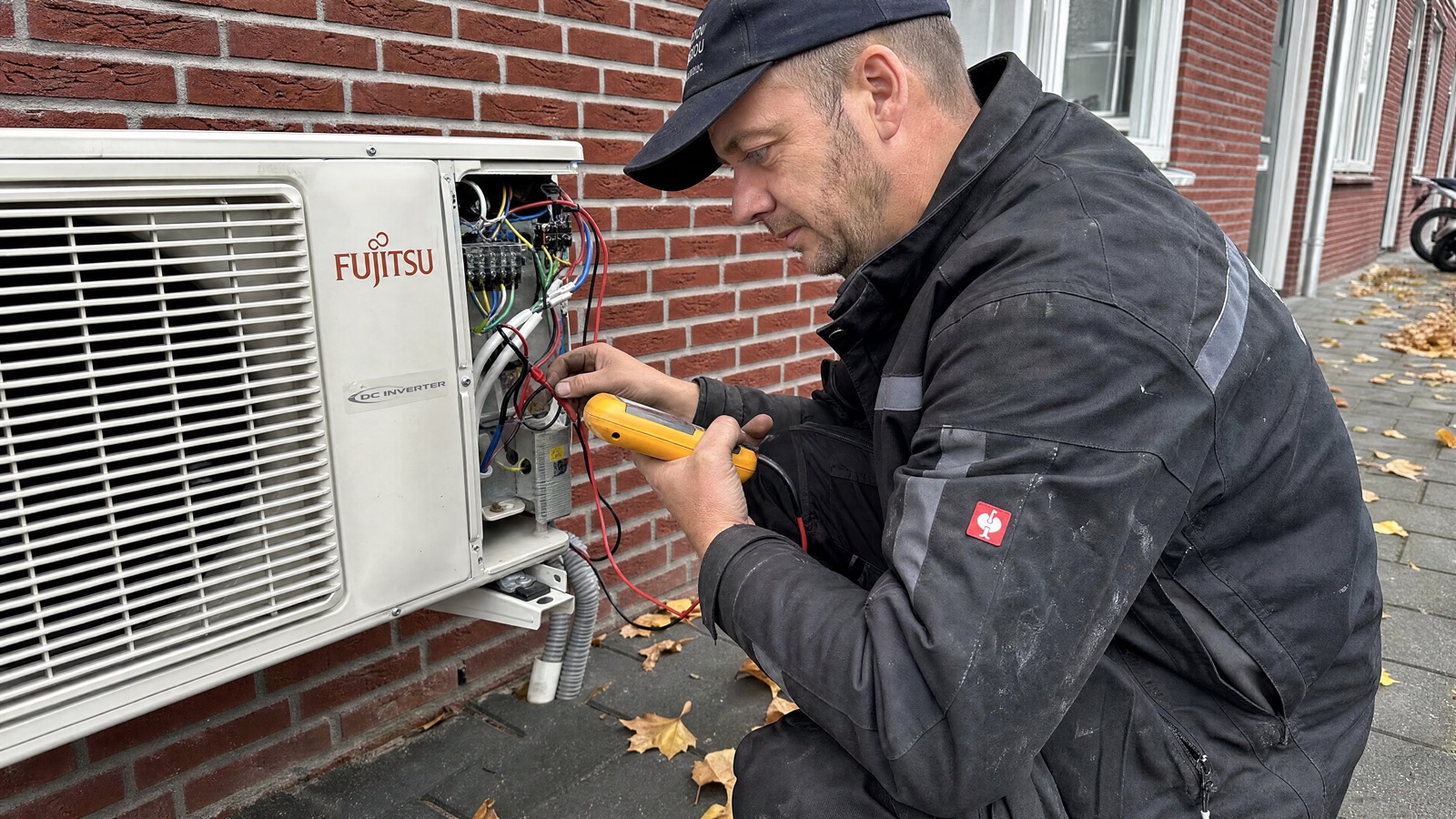 Monteur meet met een multimeter de bedrading van de buiten-unit van een airco aan een bakstenen gevel