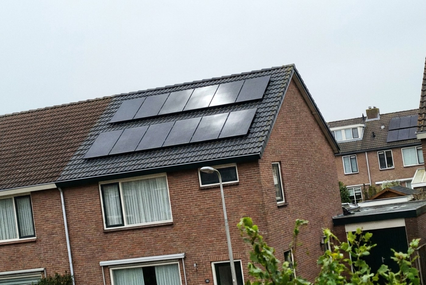 Twee rijen zwarte zonnepanelen op het grijze pannendak van een Nederlandse bakstenen woning onder een bewolkte hemel